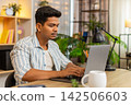 Indian man on home table typing on laptop during remote work in living room with focused expression 142506603