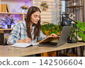 Young woman on home table taking study notes with laptop and book for focused online learning course 142506604