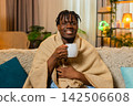 African American young man on home sofa warms hands on tea cup under blanket enjoying cozy comfort 142506608