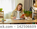 Young woman on home table gets new order alert on phone feels happy then notes details in notebook 142506609