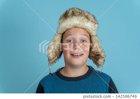 Boy smiles wearing a furry hat against a plain blue background Boy smiles wearing a furry hat against a plain blue background 142506749