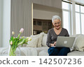 Confident stylish happy middle aged senior woman using laptop at home. Stylish older mature 60s grandmother sitting at couch looking at computer screen typing chatting reading writing email 142507236