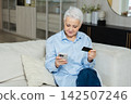Senior woman shopping online with smartphone paying with credit card at home Old lady buying on Internet enter credit card details Online shopping delivery service. Older generation modern tech usage 142507246