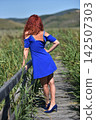 Red-haired woman in a vibrant blue dress, standing on a wooden footbridge 142507303