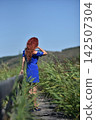Red-haired woman in a vibrant blue dress, standing on a wooden footbridge 142507304