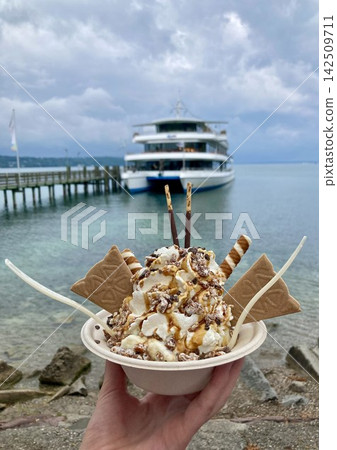 Close up view of a hand holding a plastic cup with a sweet dessert over the blurred background of a cruise ship on Lake Starnberg. Close up view of a hand holding a plastic cup with a sweet dessert over the blurred background of a cruise ship on Lake Starnberg. 142509711
