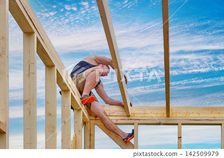 Installation of ceiling beams during construction of timber frame house, young construction worker working at height without safety harness. Installation of ceiling beams during construction of timber frame house, young construction worker working at height without safety harness. 142509979