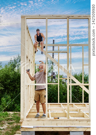 Measuring wall height with tape measure, two young construction workers work on wooden house frame outdoors. Measuring wall height with tape measure, two young construction workers work on wooden house frame outdoors. 142509980