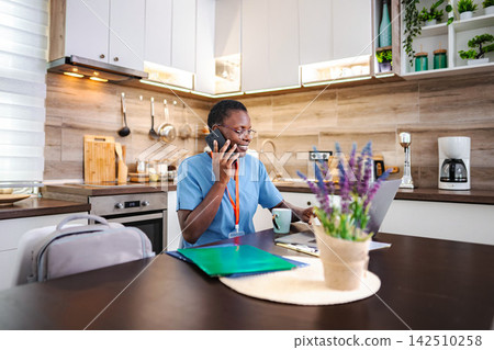 African american nurse working remotely in kitchen talking on phone African american nurse working remotely in kitchen talking on phone 142510258