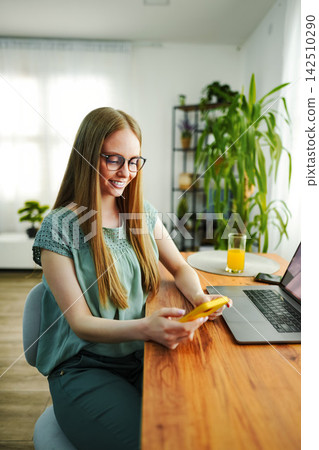 Young woman smiling using smartphone sitting at home office desk Young woman smiling using smartphone sitting at home office desk 142510290