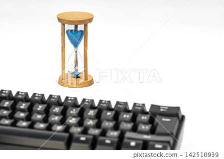 A wooden hourglass with blue sand nearly run out stands behind a black computer keyboard on a white background, symbolizing time, deadlines, and the consequences of pressing enter A wooden hourglass with blue sand nearly run out stands behind a black computer keyboard on a white background, symbolizing time, deadlines, and the consequences of pressing enter 142510939