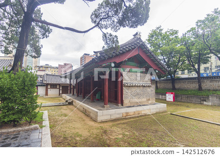 An ornate traditional Korean gate and tiled walls stand among mature trees with modern city buildings in the background. An ornate traditional Korean gate and tiled walls stand among mature trees with modern city buildings in the background. 142512676