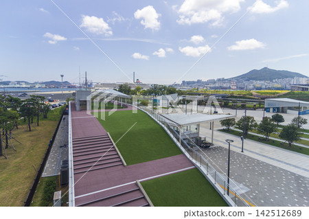 An elevated view shows a landscaped waterfront park, harbor, cruise ship, urban skyline, and mountains beneath a cloud-filled blue sky. An elevated view shows a landscaped waterfront park, harbor, cruise ship, urban skyline, and mountains beneath a cloud-filled blue sky. 142512689