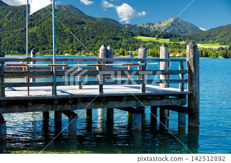 Lake pier view with wooden dock on blue alpine water with mountains background Lake pier view with wooden dock on blue alpine water with mountains background 142512892