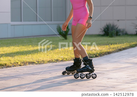 Sporty woman roller skating along a sunny park path in bright pink athletic clothing Sporty woman roller skating along a sunny park path in bright pink athletic clothing 142512978