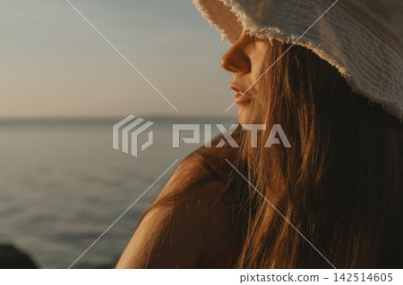 Woman in a wide brim sun hat gazes toward the calm sea at golden hour, with warm light illuminating her hair and a tranquil coastal setting creating an atmospheric summer lifestyle portrait for travel Woman in a wide brim sun hat gazes toward the calm sea at golden hour, with warm light illuminating her hair and a tranquil coastal setting creating an atmospheric summer lifestyle portrait for travel 142514605