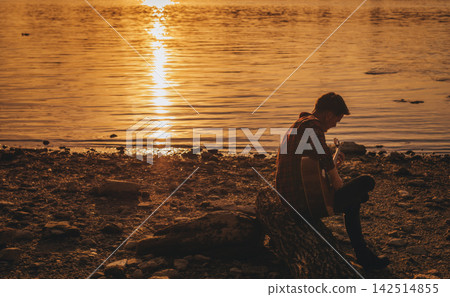 Man Playing Acoustic Guitar on River Bank at Sunset Man Playing Acoustic Guitar on River Bank at Sunset 142514855