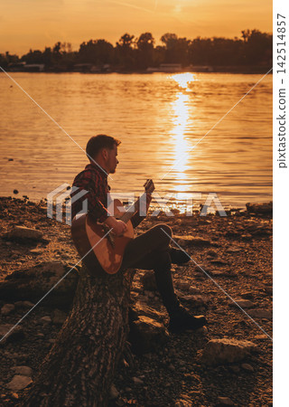 Musician Playing Acoustic Guitar on River Shore Musician Playing Acoustic Guitar on River Shore 142514857