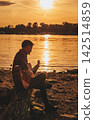 Solo Musician Playing Guitar by River at Sunset 142514859