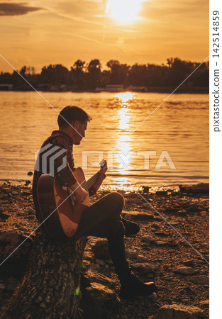 Solo Musician Playing Guitar by River at Sunset Solo Musician Playing Guitar by River at Sunset 142514859