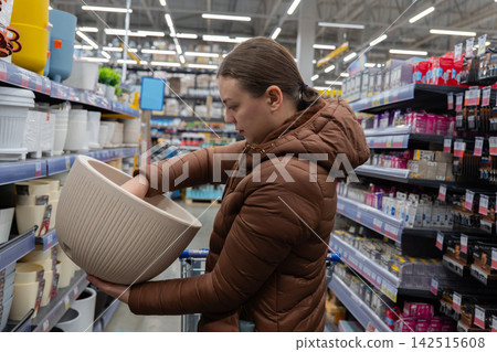 White Woman Holding Large Planter Pot In Aisle, Brown Coat, Evaluating Size And Color, Indoor Gardening Vibes, Soil And Container Sections Visible, Shopping Cart Loaded, Weekend Plant Project White Woman Holding Large Planter Pot In Aisle, Brown Coat, Evaluating Size And Color, Indoor Gardening Vibes, Soil And Container Sections Visible, Shopping Cart Loaded, Weekend Plant Project 142515608