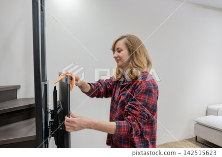 caucasian woman cleaning television screen with orange cloth in living room, focused gentle wipe removing fingerprints and dust, modern interior with neutral sofa and staircase, attentive home caucasian woman cleaning television screen with orange cloth in living room, focused gentle wipe removing fingerprints and dust, modern interior with neutral sofa and staircase, attentive home 142515623