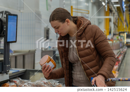 Woman examining packaged meat label closely at supermarket meat counter, checking ingredients and expiration dates, brown jacket, cart nearby, healthconscious mood, bright deli lighting, labelfocused Woman examining packaged meat label closely at supermarket meat counter, checking ingredients and expiration dates, brown jacket, cart nearby, healthconscious mood, bright deli lighting, labelfocused 142515624