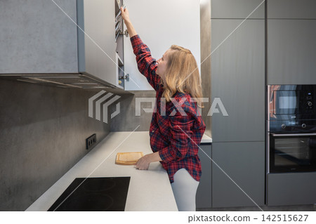 Woman reaching upper cabinet in kitchen, grabbing spice jar while prepping vegetables on wooden board, wearing red flannel shirt and white leggings, standing next to induction stove and grey 142515627