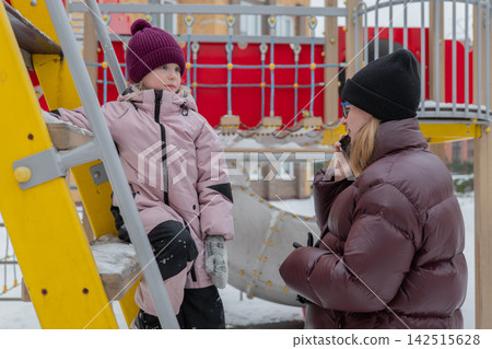 A mother and daughter are standing on the snow-covered stairs of a playground, wearing down jackets and knitted hats. The mother is holding a phone and talking animatedly. 142515628