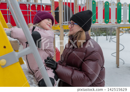 A caring adult supports a child on icy stairs, a caring caregiver helps a young girl on icy stairs outdoors, a caring adult maintains a child's balance on a slippery outdoor climbing wall. 142515629