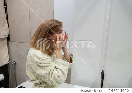 Woman in bathrobe applying moisturizer at bathroom mirror, busy professional performing evening skincare ritual after work, contemplative expression, gentle facial massage with cream, soft lighting Woman in bathrobe applying moisturizer at bathroom mirror, busy professional performing evening skincare ritual after work, contemplative expression, gentle facial massage with cream, soft lighting 142515630