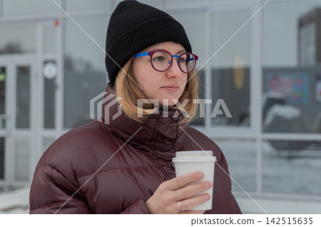 Woman holding coffee cup outdoors, contemplative commuter in maroon puffer jacket and black beanie, round glasses framing calm gaze, snowdusted urban backdrop, muted winter light, soft breath visible Woman holding coffee cup outdoors, contemplative commuter in maroon puffer jacket and black beanie, round glasses framing calm gaze, snowdusted urban backdrop, muted winter light, soft breath visible 142515635