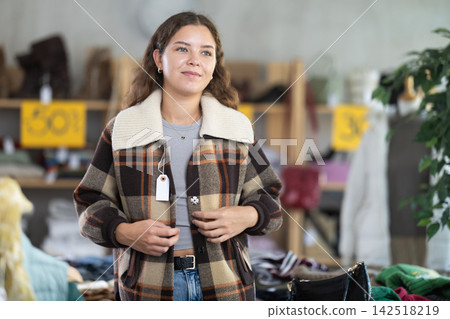 During November sales, young woman choose and try on checkered coat-jacket, view reflection in mirror. During November sales, young woman choose and try on checkered coat-jacket, view reflection in mirror. 142518219