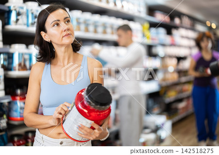 Focused woman choosing protein powder jar in sports nutrition store Focused woman choosing protein powder jar in sports nutrition store 142518275