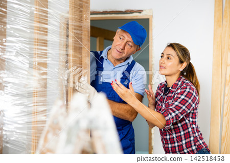 Female in casual clothes, talking with the middle-aged craftsman in uniform while he is holding a wooden door in the house Female in casual clothes, talking with the middle-aged craftsman in uniform while he is holding a wooden door in the house 142518458