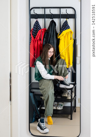 Teenage girl tying school shoes sitting on entryway bench under rack of yellow and red coats. Focused expression adjusting laces with backpack at feet in soft morning light. Gen Z student back to 142518591