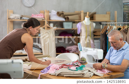 Senior man employee sewing, stitch clothes, girl apprentice draw dress pattern Senior man employee sewing, stitch clothes, girl apprentice draw dress pattern 142518691