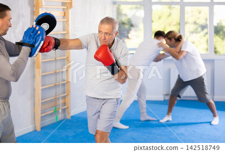 Coach and elderly man pupil are engaged during training, boxing classes. Male trainer holds makiwara and helps student to work out force of blow, to box quickly and effectively Coach and elderly man pupil are engaged during training, boxing classes. Male trainer holds makiwara and helps student to work out force of blow, to box quickly and effectively 142518749