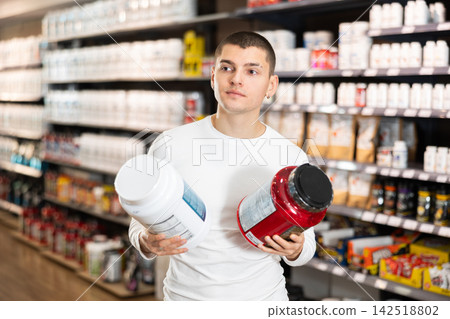 Young guy choosing protein powder jar in sports nutrition store Young guy choosing protein powder jar in sports nutrition store 142518802