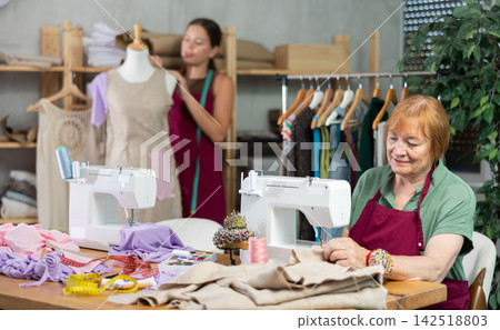 Mature dressmaker sewing clothes against the background of assistant and mannequin Mature dressmaker sewing clothes against the background of assistant and mannequin 142518803