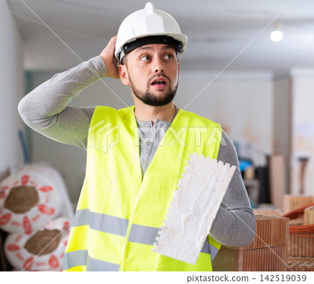 Frustrated worker in yellow vest and hard hat is dissatisfied with the way walls are plastered Frustrated worker in yellow vest and hard hat is dissatisfied with the way walls are plastered 142519039
