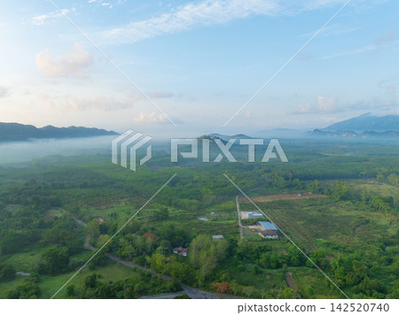 Aerial view of lush tropical countryside covered by morning mist, with green forest, rural homes and distant mountains beneath blue sky, revealing a peaceful landscape during the rainy season. Aerial view of lush tropical countryside covered by morning mist, with green forest, rural homes and distant mountains beneath blue sky, revealing a peaceful landscape during the rainy season. 142520740