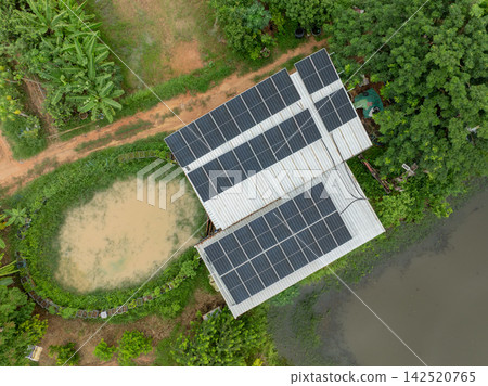 Top down drone view of rooftop solar panels on a rural farm building beside irrigation ponds and tropical crops during rainy season. Renewable electricity for sustainable agriculture and clean energy. 142520765