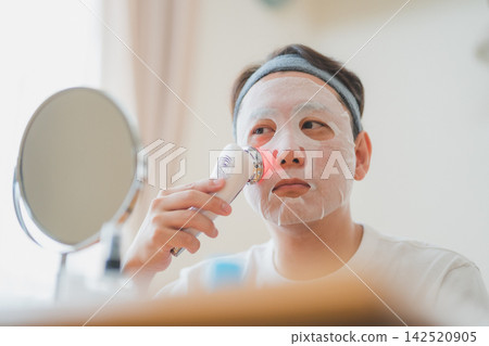 A 40-year-old man's men's beauty routine: applying a facial device to his cheeks over a face mask for intensive care. A 40-year-old man's men's beauty routine: applying a facial device to his cheeks over a face mask for intensive care. 142520905