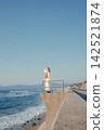 A woman with a camera stands on a breakwater with the blue sky and sea as a backdrop. 142521874