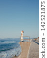 A woman with a camera stands on a breakwater with the blue sky and sea as a backdrop. 142521875