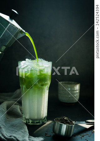 Poured Matcha green tea with Italian Moka pot pouring into a cup with iced of milk and powdered green tea on spoon on black background. copy space for your text Poured Matcha green tea with Italian Moka pot pouring into a cup with iced of milk and powdered green tea on spoon on black background. copy space for your text 142524394