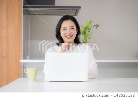A smiling woman in her 50s uses her computer in her home kitchen, enjoying a lifestyle that involves working from home using online services. A smiling woman in her 50s uses her computer in her home kitchen, enjoying a lifestyle that involves working from home using online services. 142525158