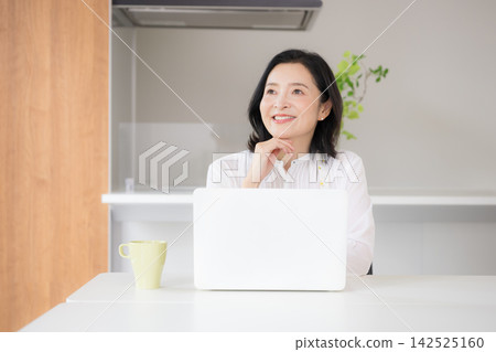 一位微笑的五十多歲女性在家裡的廚房裡用電腦打字,享受在家工作的便利。 一位微笑的五十多歲女性在家裡的廚房裡用電腦打字,享受在家工作的便利。 142525160
