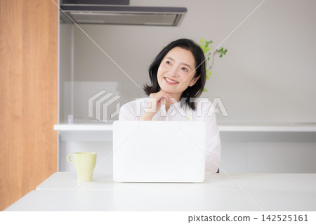 一位微笑的五十多歲女性在家裡的廚房裡用電腦打字,享受在家工作的便利。 一位微笑的五十多歲女性在家裡的廚房裡用電腦打字,享受在家工作的便利。 142525161